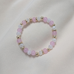 Puede incluir: Una pulsera circular de cuentas que presenta una alternancia de cuentas lisas de color rosa claro, cuentas blancas transl&uacute;cidas facetadas y peque&ntilde;os espaciadores esf&eacute;ricos met&aacute;licos de tono dorado dispuestos en un patr&oacute;n repetitivo.