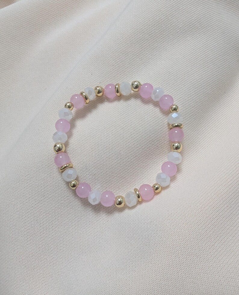 Puede incluir: Una pulsera circular de cuentas que presenta una alternancia de cuentas lisas de color rosa claro, cuentas blancas transl&uacute;cidas facetadas y peque&ntilde;os espaciadores esf&eacute;ricos met&aacute;licos de tono dorado dispuestos en un patr&oacute;n repetitivo.