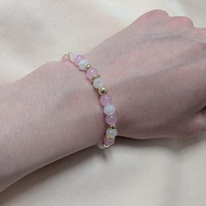 Puede incluir: Una pulsera de cuentas que presenta una alternancia de cuentas redondas rosas, cuentas blancas transl&uacute;cidas y peque&ntilde;as cuentas espaciadoras met&aacute;licas de tono dorado. La pulsera est&aacute; hecha con un cord&oacute;n el&aacute;stico flexible.