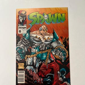 May include: A Spawn comic book issue number 6 with a cover illustration featuring a large, muscular cyborg character with a metallic chest piece and gold chains, standing over a red-cloaked figure. The title Spawn is printed in green letters.