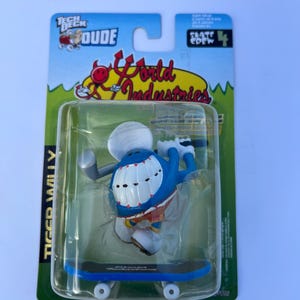 Peut inclure: Une figurine Tech Deck Dude nommée Tiger Willy, emballée sous blister. Le personnage bleu porte une casquette blanche, a de grandes dents blanches et est placé sur un skateboard bleu. L'emballage présente le logo et le texte World Industries.