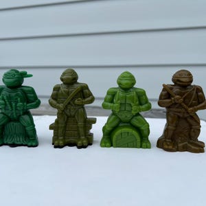May include: Four small plastic Teenage Mutant Ninja Turtles figurines standing in a row. The first is bright green, the second is olive green, the third is lime green, and the fourth is brown. Each figure is posed differently on a base.
