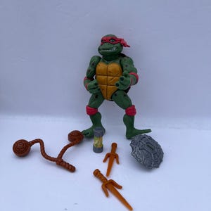 May include: A Teenage Mutant Ninja Turtle action figure, Raphael, in a fighting stance. The green figure has a red mask, orange shell, and various accessories, including sai, a weapon, and a turtle shell.