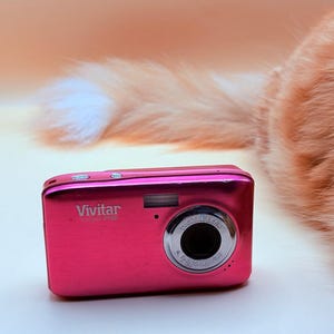 May include: A bright pink Vivitar digital camera with a silver lens and flash. The camera has the words "Vivitar" and "DIGITAL LENS" printed on it. The camera is on a white surface.