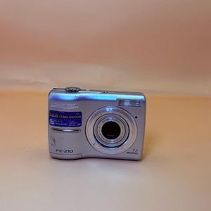 May include: A silver Olympus FE-210 digital camera featuring a 7.1 megapixel lens, a 2.5 inch LCD screen indicator, and digital image stabilisation technology. The front of the camera displays the model name and technical specifications.