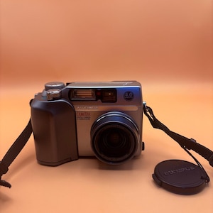 May include: Silver and black Olympus C-4000 Zoom digital camera with a lens cap and strap. The camera has a 4.0 megapixel resolution and the brand name is visible on the front.