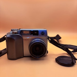 May include: A silver and gray Olympus Camedia digital camera with a black lens and a black strap. The camera has a 4.0 marking and the lens cap is next to the camera.
