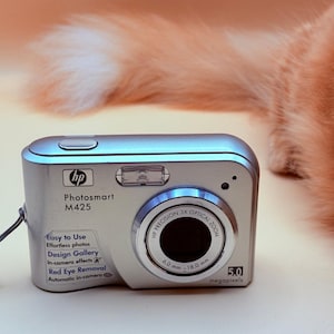 May include: Silver HP Photosmart M425 digital camera with a 3x optical zoom lens. Features include red-eye removal and in-camera effects. The camera has a 5.0 megapixel resolution.