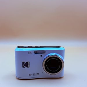 May include: A light blue Kodak digital camera with a silver lens and black buttons. The camera has the text "4X WIDE", "FULL HD", and "KODAK" printed on it. The camera is a compact size.