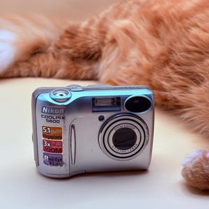 May include: Silver Nikon COOLPIX 5600 digital camera with a 5.1-megapixel resolution and 3x optical zoom. The camera features a zoom Nikkor lens and red-eye fix. The camera is on a light beige surface.