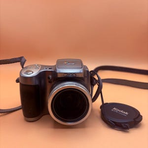 May include: A silver and black Kodak DX Zoom digital camera with a lens cap. The camera has a black hand grip and a black strap. The lens is surrounded by a silver ring. The lens cap has the Kodak DX Zoom logo.
