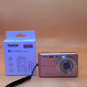 May include: A pink Casio Exilim 7.2 megapixel digital camera with a black wrist strap, positioned next to a white Kastar Slim-CNP20 charger box for camera and camcorder batteries, featuring icons for compatibility and high-speed charging.