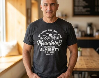 Può includere: Una t-shirt a maniche corte grigio antracite con testo serigrafato in bianco che recita I WORSHIP THE ONE WHO FORMED THE MOUNTAINS THE LORD GOD ALMIGHTY IS HIS NAME AMOS 4:13, accompagnata da un'illustrazione grafica di vette montuose.