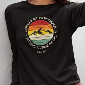 以下が含まれることがあります： 黒の長袖Tシャツ。円形のグラフィックには山の風景と「PERHAPS YOU WERE CREATED FOR SUCH A TIME AS THIS」と「Esther 4:14」の文字が描かれています。 レトロな配色です。