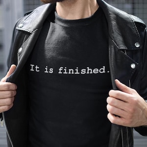 May include: Black crew neck t-shirt with the white text "It is finished." The shirt is worn under a black leather jacket. The image is a medium shot, focusing on the shirt and upper body.