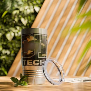 May include: A stainless steel tumbler with a camouflage design and the word "TECH" printed in black. The tumbler has a clear lid and is sitting on a wooden surface. A small sprig of green leaves is in the foreground.