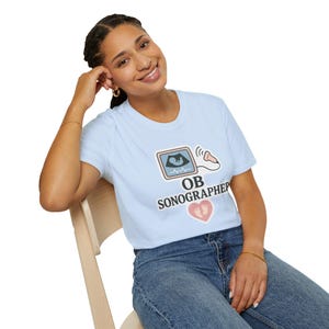 Op de afbeelding: Lichtblauw T-shirt met een afbeelding van een echo, een hartslaglijn en een roze hart met voetafdrukken. De tekst "OB SONOGRAPHER" staat eronder. Het T-shirt wordt door een persoon gedragen.