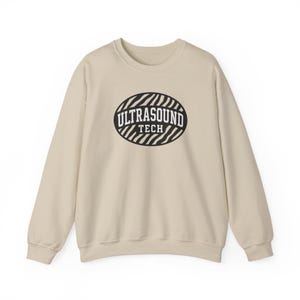 May include: A beige sweatshirt with a crew neck. The front features a black and white oval design with the words "ULTRASOUND TECH" in a bold, sans-serif font. The design is centered on the chest.