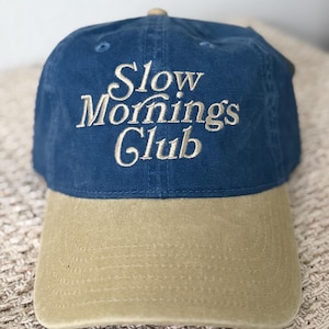 May include: A blue baseball cap with a tan brim. The phrase "Slow Mornings Club" is embroidered in a cream-colored script across the front of the cap. The cap has a curved brim and a button on top.