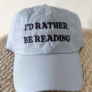 May include: A light blue baseball cap with the embroidered text "I'D RATHER BE READING" in black. The cap has a curved brim and a six-panel design, suitable for casual wear.