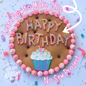 Pode incluir: Um biscoito de feltro marrom decorado com glacê rosa e branco, granulado e as palavras "HAPPY BIRTHDAY". O biscoito é cercado por mini marshmallows rosa e branco. O texto "Learn How to Make This No Sew Project" está escrito em letras rosa sobre um fundo branco.