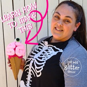 May include: A pink and white brain-shaped ice cream cone with a googly eye on top. The cone is held by a person wearing a grey shirt with a black skeleton design and the text "just add glitter decor".