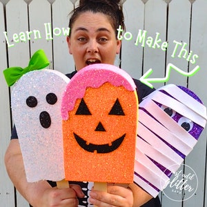 May include: Three popsicle-shaped decorations featuring a white ghost with a green bow, an orange jack-o'-lantern with pink icing, and a purple mummy with white bandages. The text "Learn How to Make This" and "just add glitter SATX" are visible.