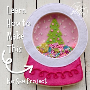 May include: A handmade, no-sew snow globe craft project. The snow globe features a pink base and a white frame. Inside, a green felt Christmas tree and colorful candies are visible. The text "Learn How to Make This" and "No Sew Project" are also displayed.