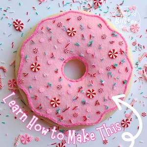May include: A pink felt donut with white frosting and red and white candy sprinkles. The donut is surrounded by white sprinkles and candy canes. The text "Learn How to Make This" is written in white on a white background.