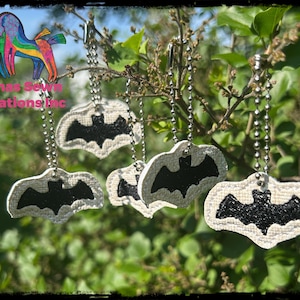 Könnte beinhalten: Fledermausförmige Schlüsselanhänger hängen an einem Ast. Jeder Schlüsselanhänger zeigt ein schwarzes Fledermausdesign auf cremefarbenem Hintergrund, befestigt an einer silbernen Kugelkette. Der Text "Llamas Sewn Creations Inc." ist enthalten.