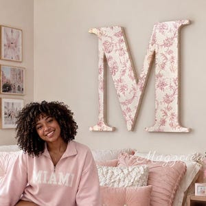 May include: A light pink quarter-zip sweatshirt with the word MIAMI printed in white across the chest. The garment features long sleeves and a high collar, worn over a bed with a textured pink comforter, matching pillows, and a pink throw blanket.