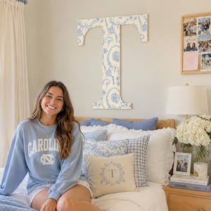 May include: A large wooden letter T wall decoration featuring a blue and white floral pattern. The letter is mounted on a wall above a bed, which is styled with various pillows, including a small rectangular cushion with a blue letter T design.