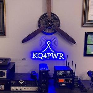Può includere: Un cartello al neon blu con il nominativo "KQ4PWR" e una grafica di antenna stilizzata. Il cartello è montato su una parete con un'elica di legno dietro. Ci sono diversi ricetrasmettitori radio e altri dispositivi elettronici su un tavolo sotto il cartello.