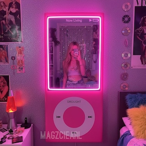 Puede incluir: Un espejo de pared rosa con forma de iPod, con un marco rosa neón. El espejo refleja a una persona haciéndose un selfie. La palabra "Drought" está impresa en la parte inferior, con "Now Living" en la parte superior.