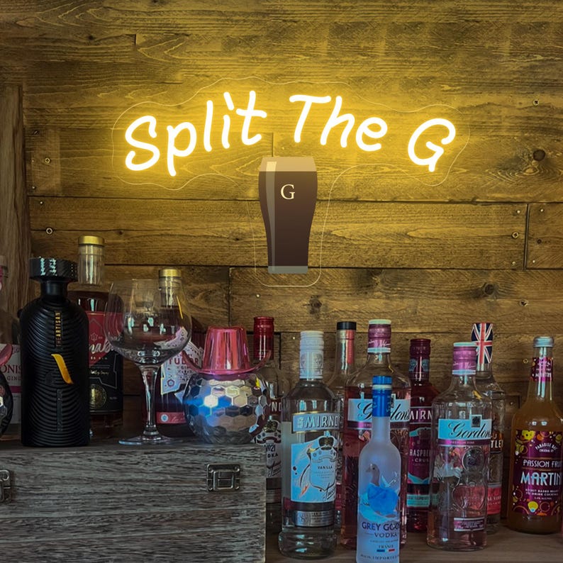 Pode incluir: Uma placa de n&eacute;on que diz "Split The G" acima de um bar com v&aacute;rias garrafas de licor. A placa &eacute; amarela e apresenta um gr&aacute;fico de caneca. O bar tem um fundo de madeira.