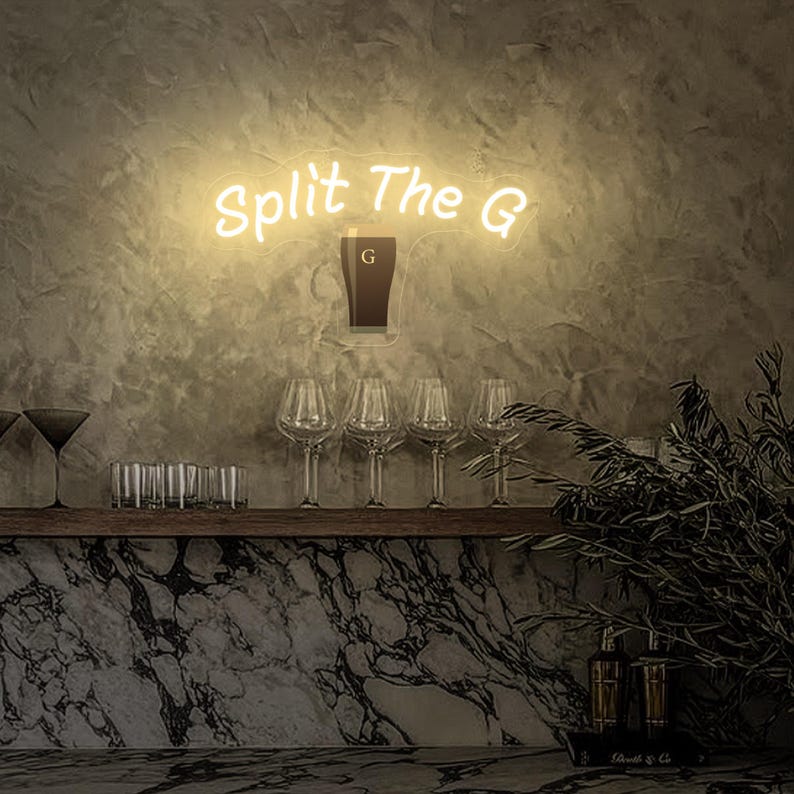 Pode incluir: Uma placa de n&eacute;on que diz "Split The G" em fonte cursiva, com um gr&aacute;fico de um copo de cerveja. Abaixo da placa est&atilde;o quatro ta&ccedil;as de vinho e uma prateleira com outros copos. O fundo &eacute; uma parede texturizada.