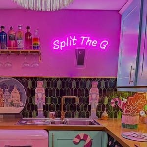 Pode incluir: Uma cena de cozinha com uma placa de n&eacute;on que diz "Split The G" acima de uma pia. A cozinha tem um esquema de cores rosa e verde, com garrafas e copos em uma prateleira. Quebra-nozes decorativos e uma geladeira rosa tamb&eacute;m s&atilde;o vis&iacute;veis.