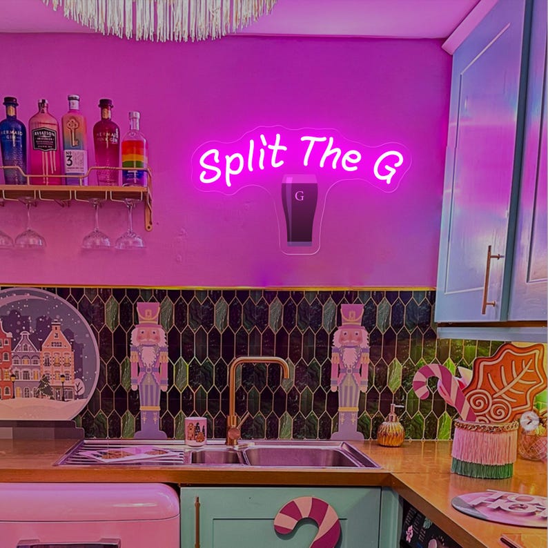 Pode incluir: Uma cena de cozinha com uma placa de n&eacute;on que diz "Split The G" acima de uma pia. A cozinha tem um esquema de cores rosa e verde, com garrafas e copos em uma prateleira. Quebra-nozes decorativos e uma geladeira rosa tamb&eacute;m s&atilde;o vis&iacute;veis.