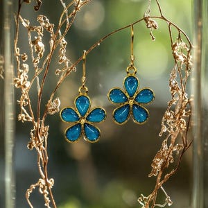 May include: A pair of flower-shaped earrings with a gold-colored frame and bright blue petals. The earrings are displayed inside a glass cloche with dried plants and moss.