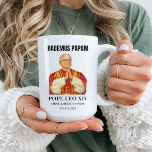 May include: White ceramic mug featuring an image of Pope Leo XIV with the text "HABEMUS PAPAM" and "First American Pope, May 8, 2025". The mug has a large handle and is held by a person.