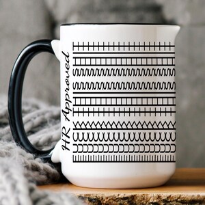 Può includere: Tazza in ceramica bianca con manico e bordo neri, con un motivo geometrico nero e la scritta "HR Approved" in nero. La tazza è esposta su una superficie di legno con una coperta di maglia grigia sullo sfondo.