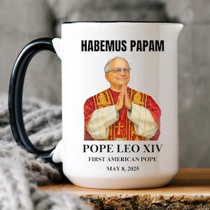 May include: White ceramic mug with a black handle and rim. The mug features an image of Pope Leo XIV with the text "HABEMUS PAPAM" above and "POPE LEO XIV FIRST AMERICAN POPE MAY 8, 2025" below the image.