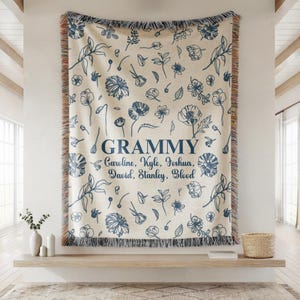 May include: A cream-colored throw blanket with a blue floral pattern and the word "Grammy" in navy blue. The names Caroline, Kyle, Joshua, David, Stanley, and Blood are below the word "Grammy". The blanket has a fringed edge.