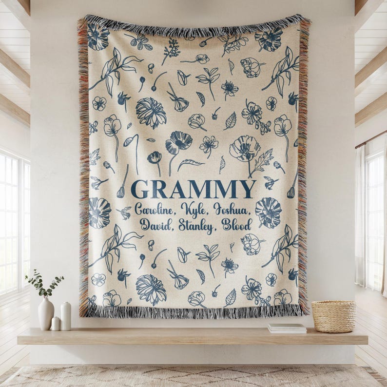 May include: A cream-colored throw blanket with a blue floral pattern and the word "Grammy" in navy blue. The names Caroline, Kyle, Joshua, David, Stanley, and Blood are below the word "Grammy". The blanket has a fringed edge.