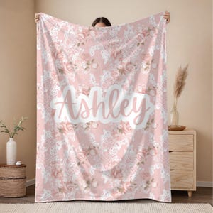 May include: A pink blanket with a floral and lace pattern. The blanket features the name "Ashley" in a white, cursive font. The design includes pink roses and white lace details, creating a soft and feminine aesthetic.