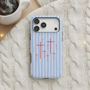 May include: A light blue and white striped phone case featuring a pattern of red crosses. The case is designed to fit a smartphone and is shown on a white cable knit sweater.
