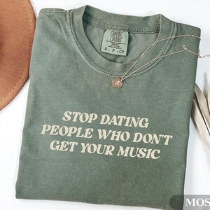 May include: Moss green t-shirt with the text "STOP DATING PEOPLE WHO DON'T GET YOUR MUSIC" in a white font. A gold necklace with a pendant rests on the shirt. The shirt is folded and placed on a white surface.