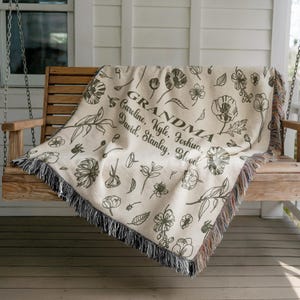 May include: Cream-colored throw blanket with a floral pattern and the word "GRANDMA" in the center, along with several names. The blanket has a fringe border and is draped over a wooden swing.