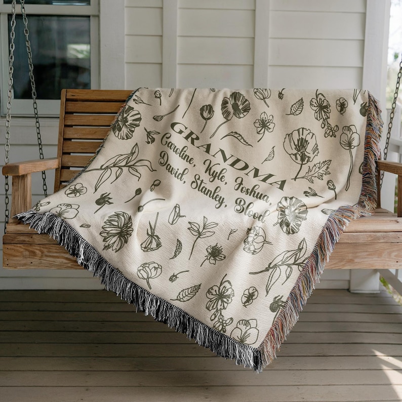 May include: Cream-colored throw blanket with a floral pattern and the word "GRANDMA" in the center, along with several names. The blanket has a fringe border and is draped over a wooden swing.