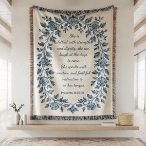 May include: Cream-colored woven throw blanket with a blue floral design and text from Proverbs 31:25-26. The text is framed by a wreath of blue flowers and leaves. The blanket has a fringed edge.